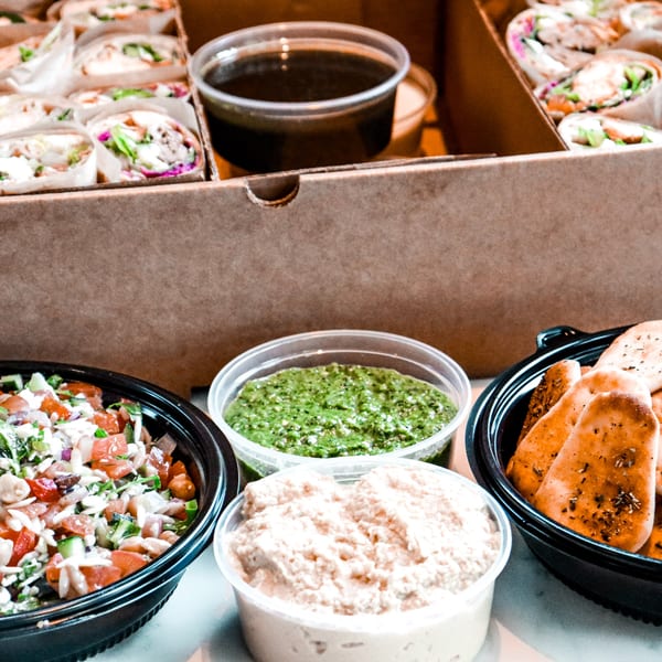 Tazā Mediterranean catering box with wraps, salad, pita bread, hummus, and green sauce.