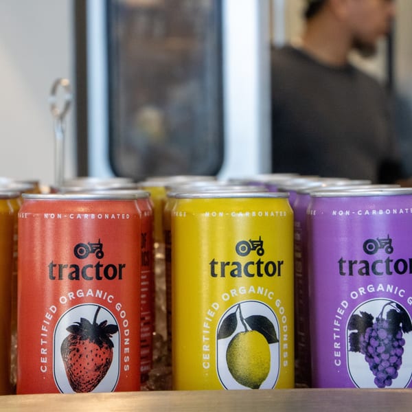 Three colorful Tractor Organic Goodness cans: red strawberry, yellow lemon, and purple grape.