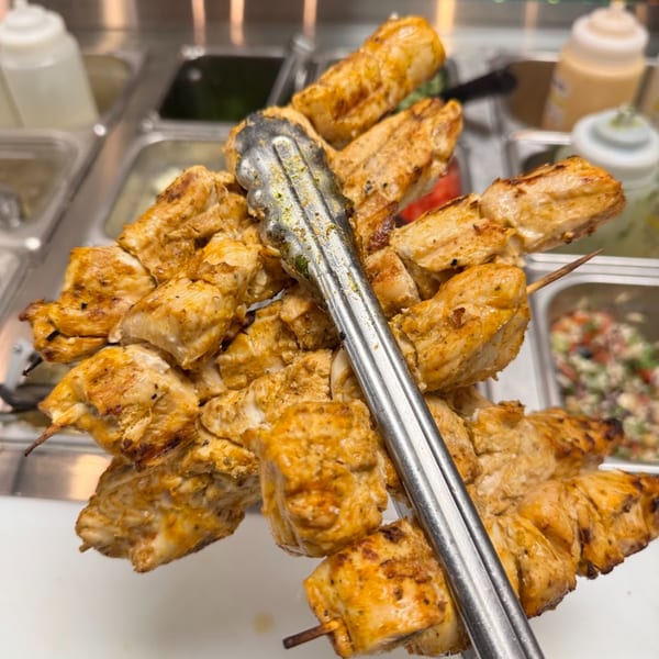Grilled chicken shish tawook skewers held by tongs, with salad bar containers in the background.