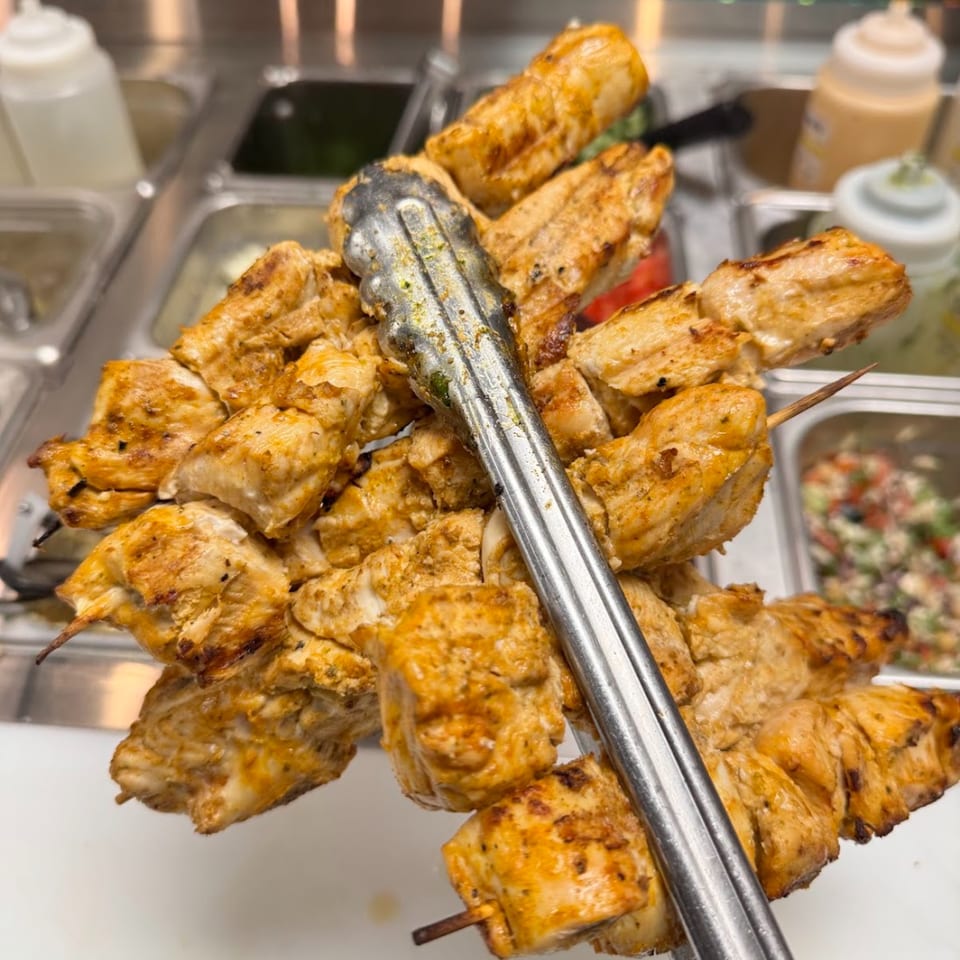 Grilled chicken shish tawook skewers held by tongs, with salad bar containers in the background.
