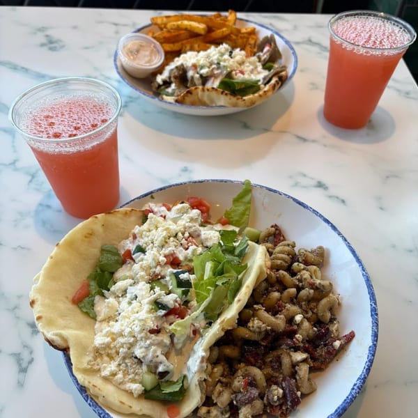 Two Tazā Mediterranean meals with gyros, fries, pasta, and two pink drinks on a marble table.