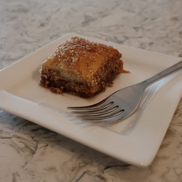 A square piece of baklava, sprinkled with nuts, on a white plate with a fork.