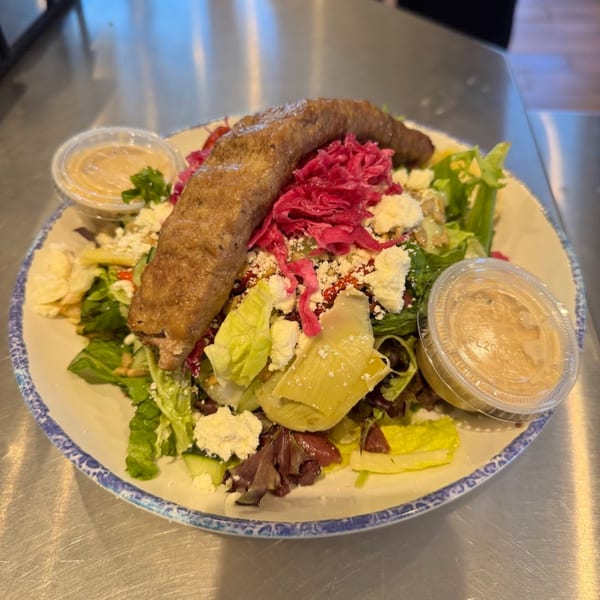 Tazā's Mediterranean salad with a long sausage, red cabbage, feta, and two dressing cups.