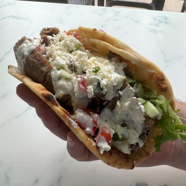 Hand holding a Tazā gyro with meat, feta, tomatoes, cucumbers, and tzatziki sauce.